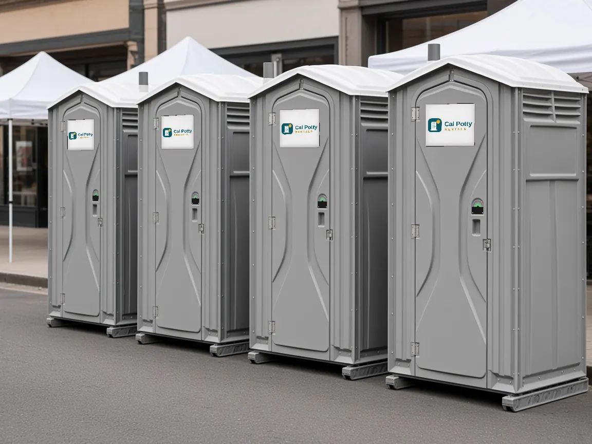 Four grey portable toilets lined up beside a paved event area with white tents