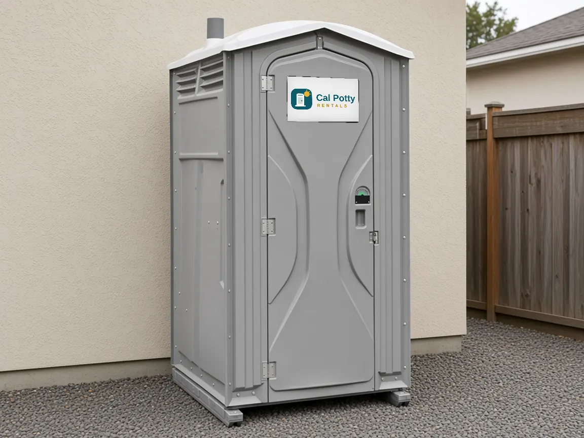 A grey standard porta potty on gravel in a side yard between a house wall and a wooden fence